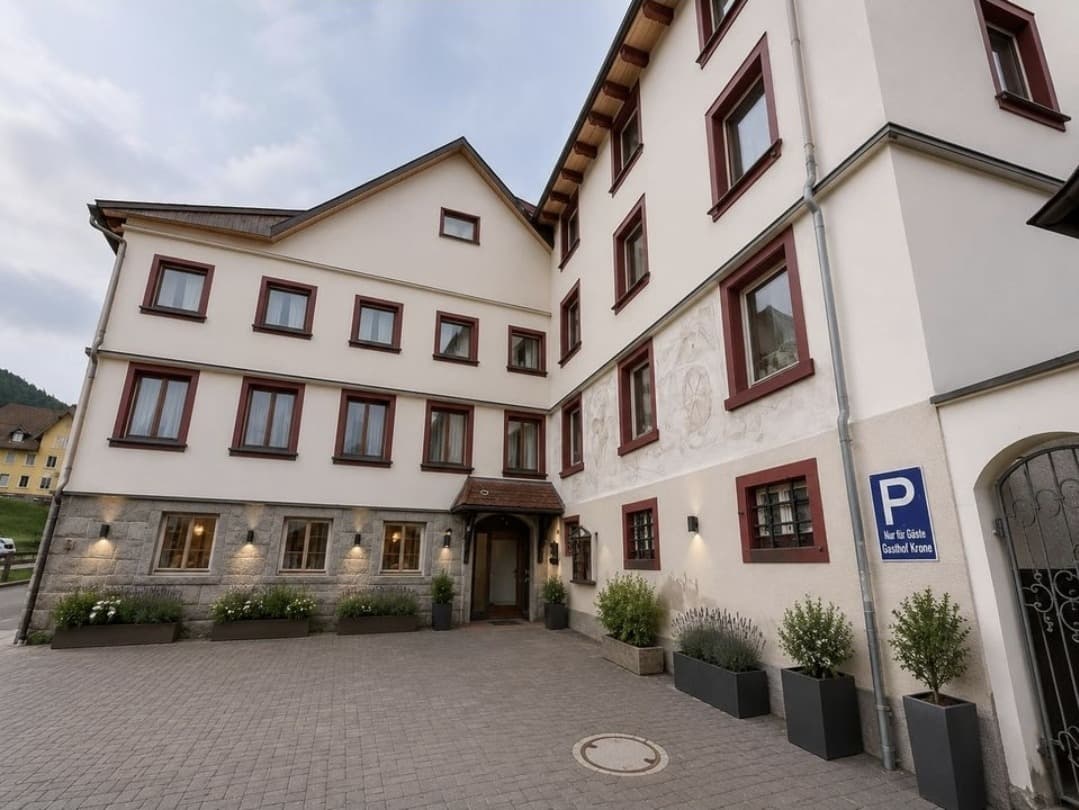 Ansicht von Hof und Eingang des Gasthof Krone nach der Renovierung: helle Fassade mit dunkelroten Fensterrahmen, gepflasterter Vorplatz mit Pflanzkübeln